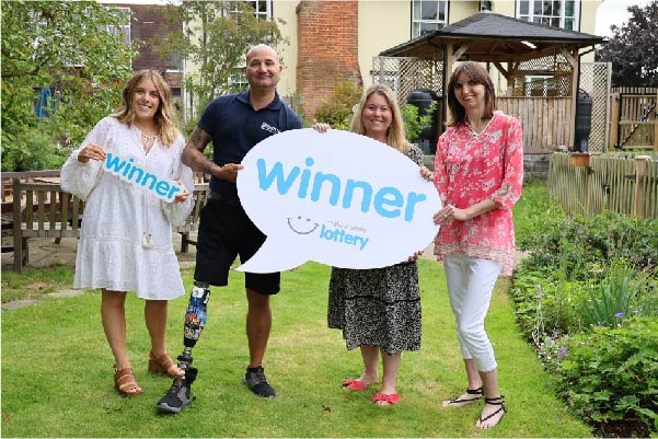 Group of young women and a limbless veteran smiling in a garden holding a make a smile lottery winner sign.