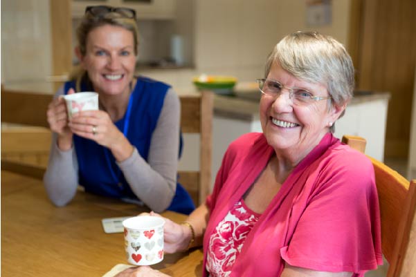 Old lady in a pink jumper with support worker sat having a cup of tea at the table